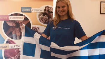 A girl holds up the Greek flag in front of a study abroad poster